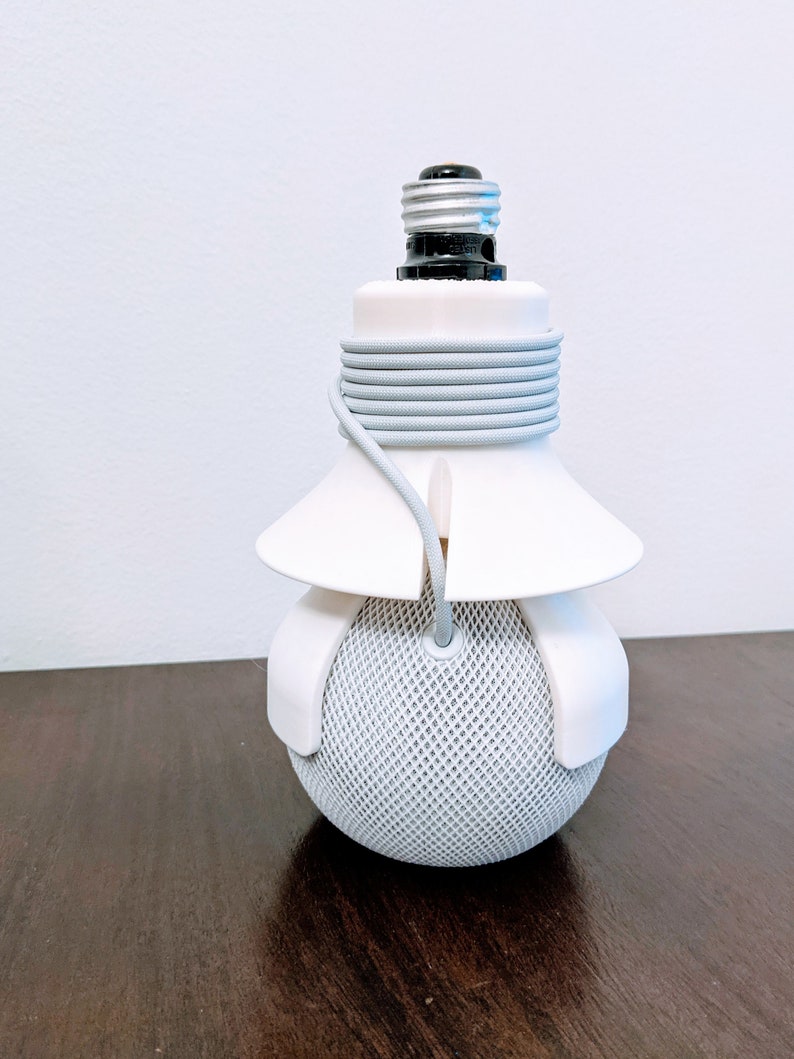 May include: A white lightbulb socket adapter with a grey cord wrapped around it. The adapter is attached to a white speaker with a grey mesh covering.