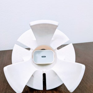 May include: White plastic phone stand with six arms, designed to hold a white wireless charging pad. The charging pad has a small black line on the front.