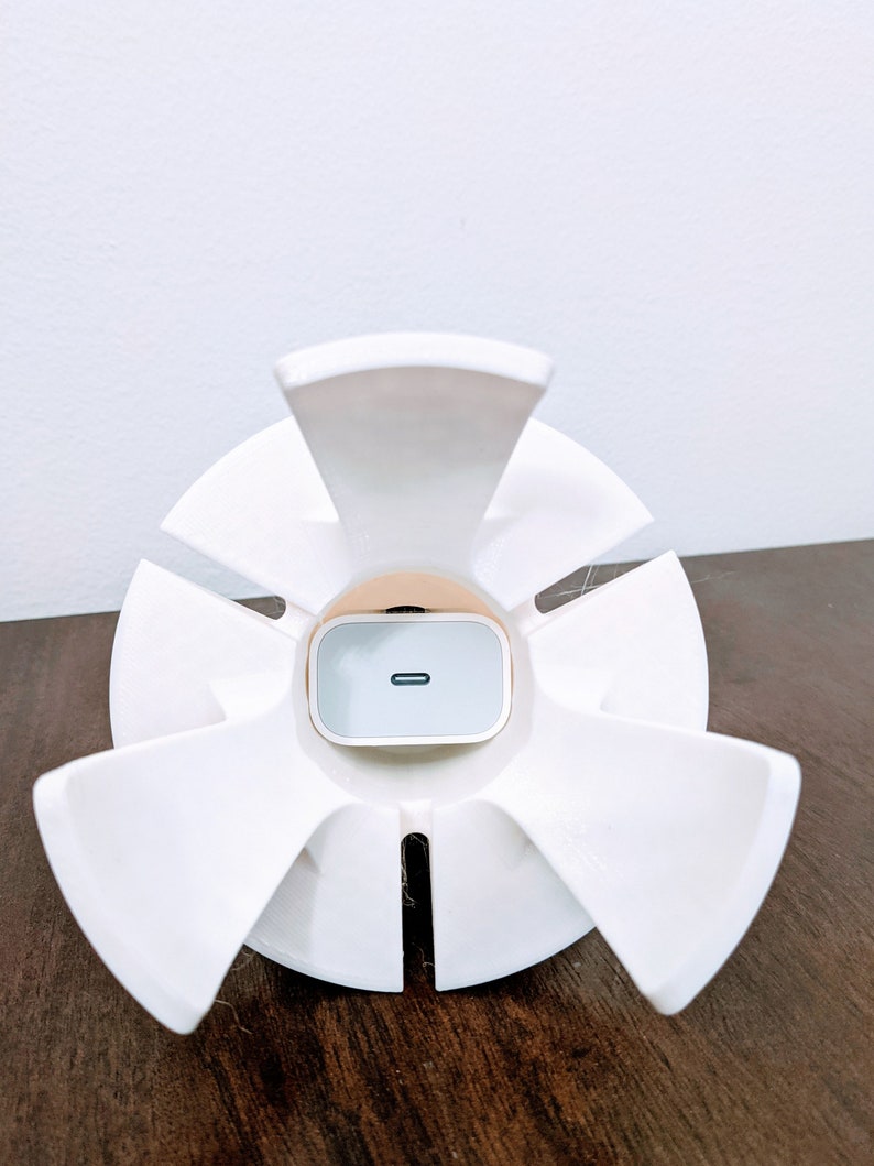May include: White plastic phone stand with six arms, designed to hold a white wireless charging pad. The charging pad has a small black line on the front.