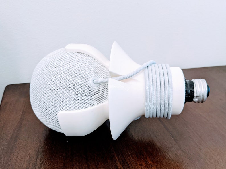 May include: White 3D printed light bulb socket adapter with a white cord wrapped around it. The adapter is designed to fit a smart speaker.