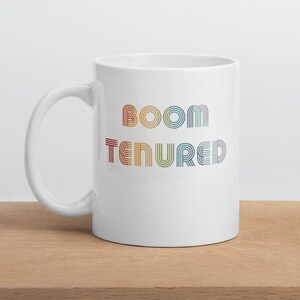 May include: White ceramic coffee mug with the text "BOOM TENURED" in rainbow colors.
