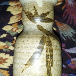Puede incluir: Un pequeño jarrón de cerámica marrón y blanco con un diseño marrón estilizado. El jarrón tiene un cuerpo redondeado y un cuello estrecho.