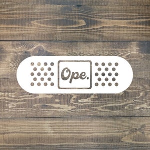 May include: White band-aid with a white square in the center that says "Ope." The band-aid is on a brown wooden background.