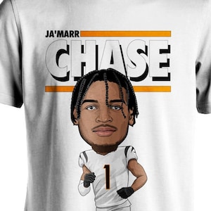 May include: White t-shirt with a cartoon illustration of Ja'Marr Chase, a football player wearing a black and orange jersey with the number 1. The text "JA'MARR CHASE" is printed in black and orange on the shirt. The text "JB STUDIO" is printed in black on the bottom of the shirt.