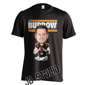 May include: Black t-shirt with a cartoon illustration of Joe Burrow, a football player, wearing a Cincinnati Bengals jersey and holding a football. The text "JOE BURROW" is printed in white on a black and orange stripe above the illustration. The text "JB STUDIO" is printed in white on a black background in the lower right corner of the shirt.