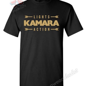 Peut inclure: T-shirt noir avec du texte doré qui dit "LIGHTS KAMARA ACTION" avec des flèches de chaque côté du texte.