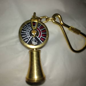 May include: A gold-colored ship's wheel keychain with a black and white dial and red markings. The dial has the words "AHEAD", "SLOW", "HALF", "FULL", "ASTERN", "SLOW", "HALF", and "FULL" printed on it. The keychain has a gold-colored ring.