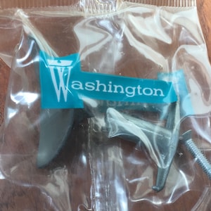 May include: A clear plastic bag containing two dark gray cabinet pulls and a silver screw. The bag has a teal and white label with the word "Washington" printed on it. The cabinet hardware is likely for furniture.