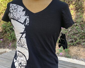 May include: A black V-neck t-shirt featuring a large, off-white graphic panel on the left side filled with various cartoon characters and the word latte. A black, skull-shaped hang tag with the brand name tokidoki is attached to the sleeve.