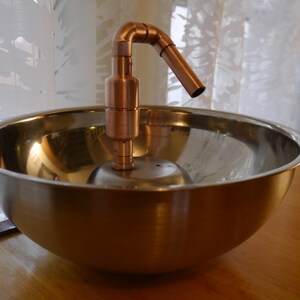 May include: A copper tap with a curved spout sits atop a stainless steel bowl. The bowl is on a wooden surface.