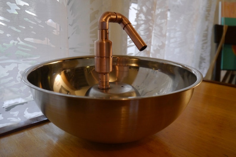 May include: A copper tap with a curved spout sits atop a stainless steel bowl. The bowl is on a wooden surface.