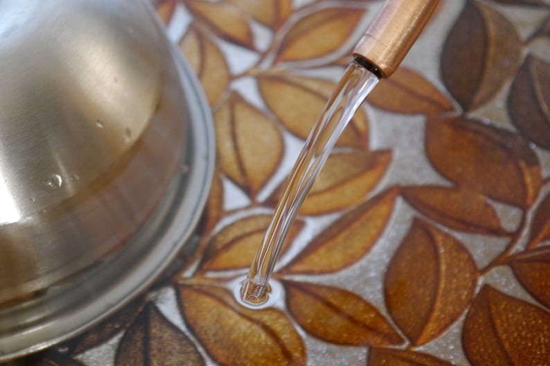 May include: A close-up of a copper tap with water flowing into a sink with a leaf pattern.