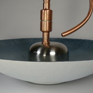 May include: A copper and stainless steel fountain with a curved spout, sitting on a white and blue ceramic bowl.