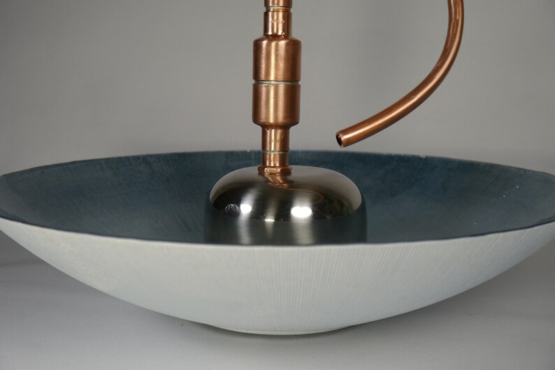 May include: A copper and stainless steel fountain with a curved spout, sitting on a white and blue ceramic bowl.