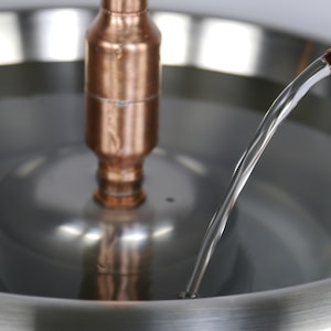 May include: A close-up of a copper pipe pouring water into a stainless steel bowl. The water is flowing in a thin stream and is creating a small ripple in the bowl.