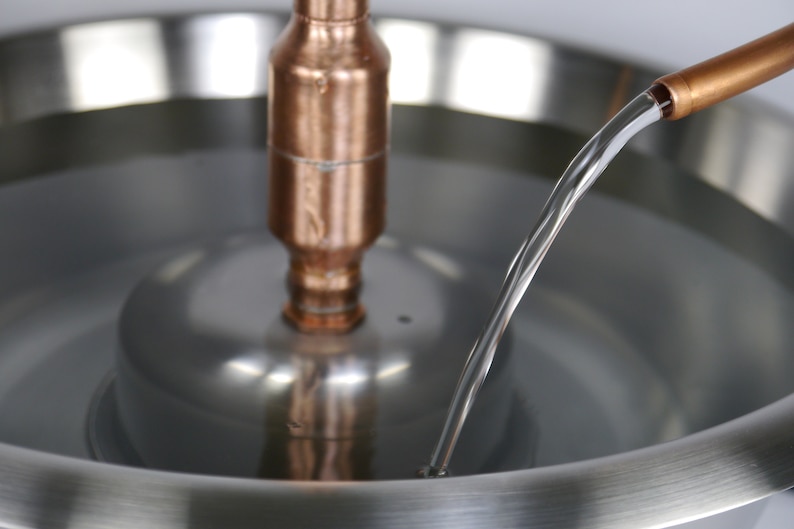 May include: A close-up of a copper pipe pouring water into a stainless steel bowl. The water is flowing in a thin stream and is creating a small ripple in the bowl.