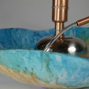 May include: A copper and stainless steel fountain with a blue and brown glass bowl. Water flows from a copper pipe into the bowl.