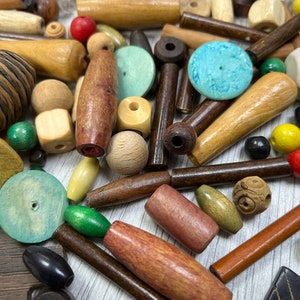 May include: A collection of wooden beads in various shapes, sizes, and colors, including brown, beige, green, blue, and red. Some beads have a natural wood finish, while others are painted or stained. The beads are scattered on a light wood surface.