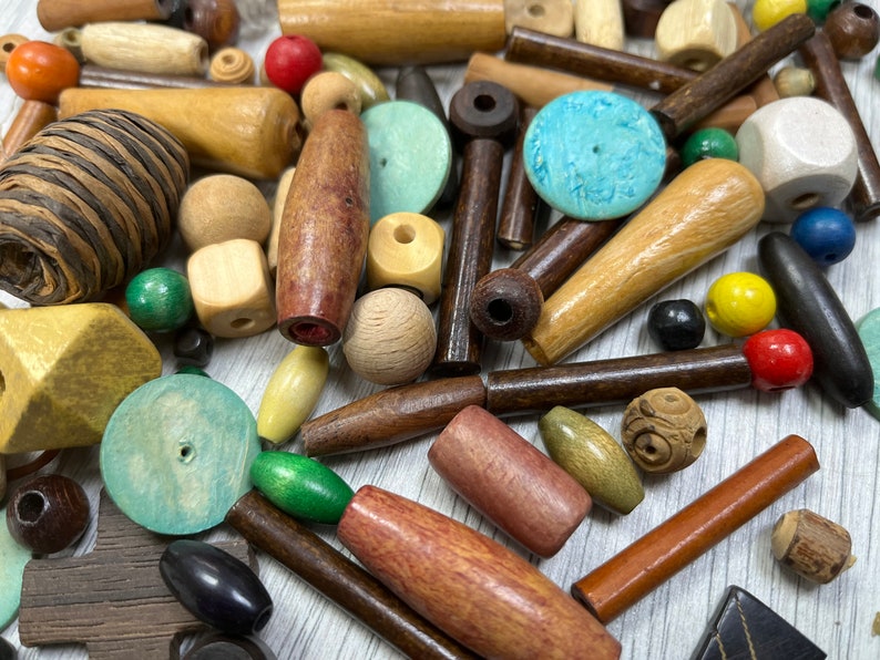 May include: A collection of wooden beads in various shapes, sizes, and colors, including brown, beige, green, blue, and red. Some beads have a natural wood finish, while others are painted or stained. The beads are scattered on a light wood surface.