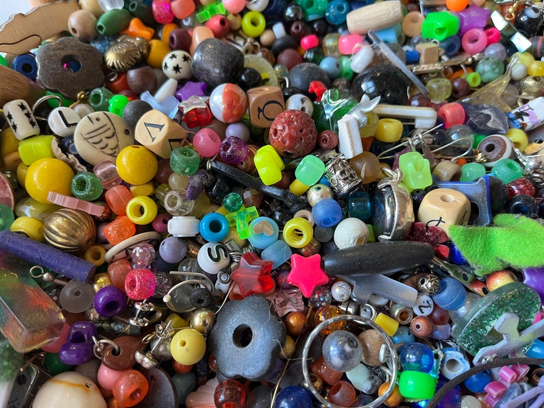 May include: A colorful assortment of beads and small decorative items. The collection includes various shapes, sizes, and colors, such as round, star, and cube-shaped beads. Materials include plastic, metal, and wood. The beads are in shades of red, yellow, green, blue, and purple.