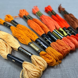 May include: A collection of embroidery floss spools in shades of orange, yellow, and brown. The spools are labelled "Fast Colours 100% Cotton".