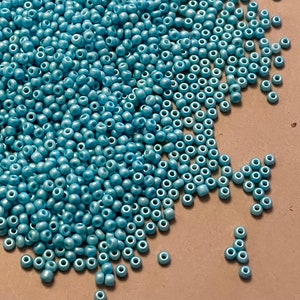 May include: A large pile of small, round, light blue seed beads. The beads have a metallic sheen and are scattered on a brown surface.