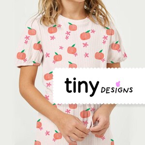Peut inclure: Un t-shirt à manches courtes rose et blanc avec un imprimé pêche et le texte "tiny DESIGNS" sur une étiquette blanche.