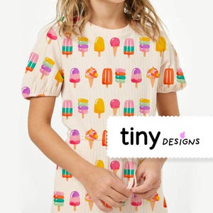 Peut inclure: Robe crème avec un motif de glaces et de cornets colorés. La robe a des manches courtes bouffantes et le texte "tiny designs" dans un rectangle blanc.