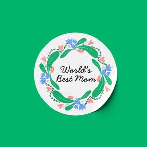 Peut inclure: Autocollant rond blanc avec une couronne de feuilles vertes et des fleurs roses et bleues. Le texte "World's Best Mom" est au centre.