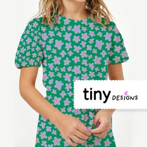 Peut inclure: Robe verte ornée d'un motif floral de fleurs lavande et orange. La robe a des manches courtes et un col rond. Le texte "tiny designs" est affiché en bas à droite de la robe.