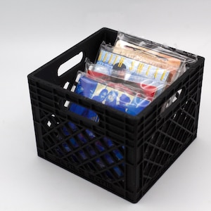 May include: A black plastic milk crate filled with several packaged items. The items are wrapped in clear plastic and appear to be various sizes and colors. The crate has a grid-like pattern and handles on the sides.