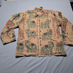 May include: A tan long-sleeve button-down shirt featuring a repeating pattern of green and dark blue elephants with red accents. The garment has a collar, two front pockets, and a textured fabric finish with decorative stitching along the edges.