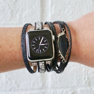 Op de afbeelding: Een zwarte Apple Watch met een zilveren wijzerplaat en een zwarte leren band. Het horloge is versierd met een gelaagde zwarte en zilveren armband met een zwarte steenaccent.