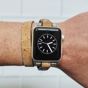 May include: A silver Apple Watch with a black face and a brown cork band. The watch is worn on a person's wrist.