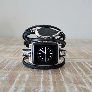 Op de afbeelding: Zwarte Apple Watch met een zwarte leren band en zilveren accenten. De band is gelaagd met meerdere wikkelingen en heeft een decoratief stenen accent.