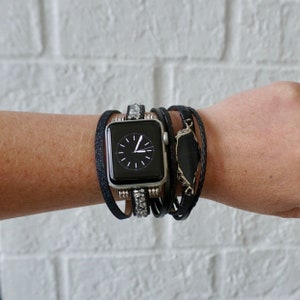 Op de afbeelding: Een zwarte Apple Watch met een zwart en zilveren bandje met strass. Het horloge wordt gedragen op een pols met meerdere zwarte leren armbanden en een zwarte stenen hanger.