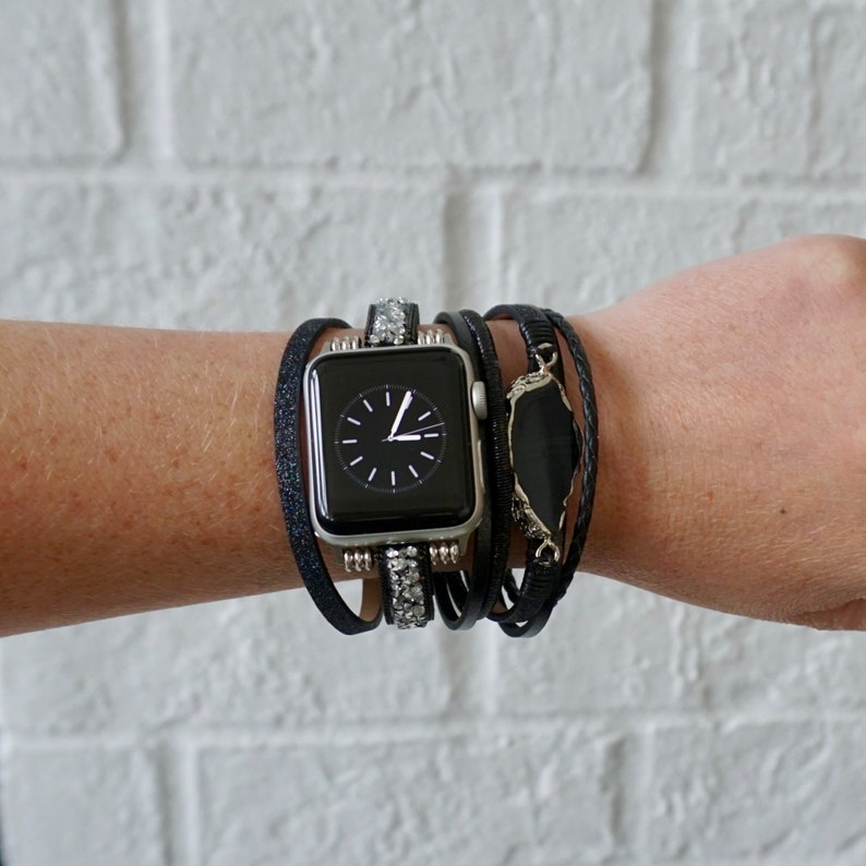Op de afbeelding: Een zwarte Apple Watch met een zwart en zilveren bandje met strass. Het horloge wordt gedragen op een pols met meerdere zwarte leren armbanden en een zwarte stenen hanger.