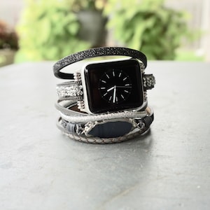 May include: A black Apple Watch with a black face and white hands is worn with a stack of five different bracelets. The bracelets are made of leather, metal, and fabric.
