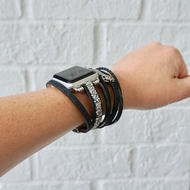 Op de afbeelding: Een zwart leren Apple Watch-bandje met zilveren accenten en strass. Het bandje is om een pols gewikkeld.