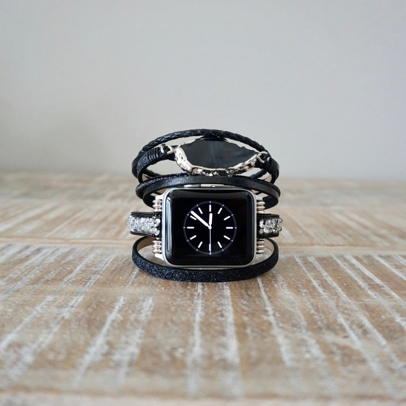 Op de afbeelding: Zwarte Apple Watch met een zwarte leren band en zilveren accenten. De band is gelaagd met meerdere wikkelingen en heeft een decoratief stenen accent.