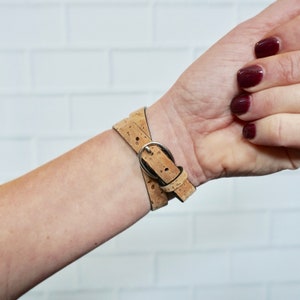 May include: A tan cork watch band with a silver buckle. The band is wrapped around a person's wrist.