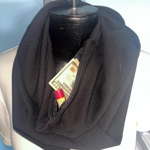 May include: A black infinity scarf with a hidden zippered pocket. The pocket contains a ten-dollar bill and a tube of lip balm. The scarf is displayed on a black mannequin head.