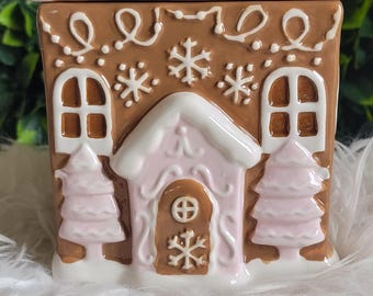 Puede incluir: Un recipiente de cerámica con forma de casa de jengibre con techo esmerilado blanco y detalles en rosa. La casa marrón presenta detalles decorativos de glaseado, ventanas y una puerta. El recipiente está diseñado para uso decorativo o funcional.
