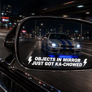 May include: A black car side mirror featuring a white vinyl decal that reads OBJECTS IN MIRROR JUST GOT KA-CHOWED with lightning bolt icons. The mirror reflects a silver sports car with blue racing stripes and glowing blue underglow on a road.