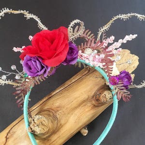 May include: A pair of turquoise headband ears with a floral arrangement featuring red, purple, and pink flowers, seashells, and brown and pink foliage.