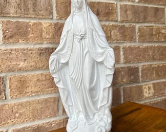 Könnte beinhalten: Eine mattweiße Figur einer stehenden Gestalt mit drapiertem Kopfschmuck und fließenden Gewändern, die Arme sind nach außen gestreckt mit den Handflächen nach vorne, platziert auf einem Sockel mit Globus und wolkenartigen Details.