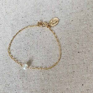 May include: A delicate gold bracelet featuring a single white pearl and a small oval charm with a religious figure. The bracelet is on a neutral-colored textured surface.