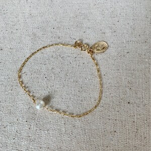 May include: A delicate gold-toned bracelet featuring a single white pearl and a small, round charm. The bracelet has a fine chain design and is displayed on a neutral linen surface.