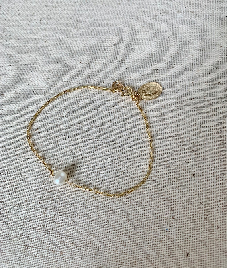 May include: A delicate gold-toned bracelet featuring a single white pearl and a small, round charm. The bracelet has a fine chain design and is displayed on a neutral linen surface.