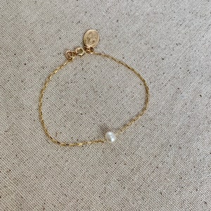 May include: Delicate gold-toned bracelet featuring a single white pearl and a small circular charm. The chain has a rectangular link design. The bracelet is displayed on a neutral-toned fabric surface.